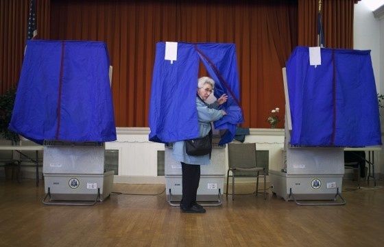 Voter entering a blue privacy booth beside ballot boxes at a polling station
